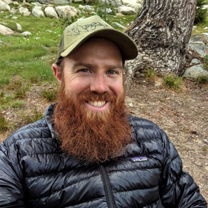 The image shows a man with a red beard and a green cap. He is wearing a black jacket and smiling at the camera. The background includes a tree and some rocks. The man appears to be outdoors, possibly in a park or natural setting.